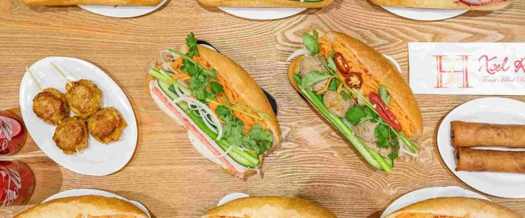 A top-down view of several banh mi sandwiches, a plate of meatballs, and two spring rolls arranged on a light wood table with a branded paper sign.