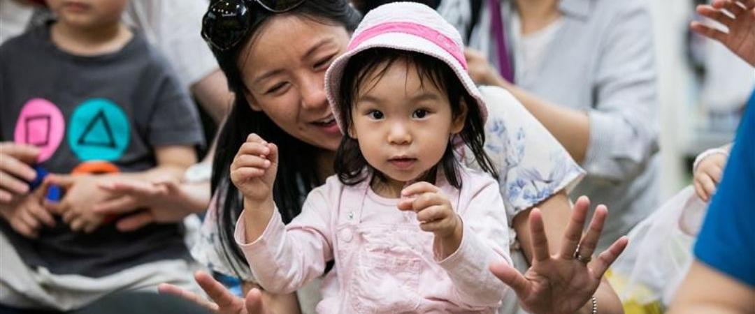 A mother holds her young daughter in a pink outfit and hat, surrounded by a diverse group of children and adults, conveying a joyful atmosphere.
