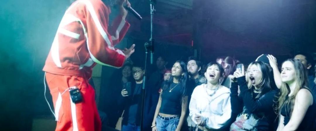 A singer in a red tracksuit performs passionately on stage with a microphone. An excited crowd, bathed in colorful stage lights, watches energetically.