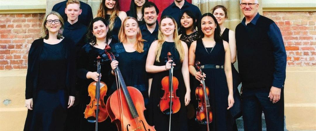 A group of musicians pose with cellos and violins in front of a red door and brick wall, wearing black formal attire, conveying a joyful and professional tone.