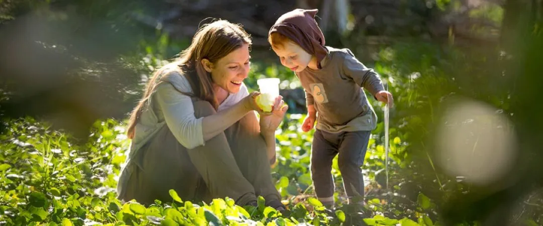 A mother crouches beside her young child in a sunlit forest, smiling as they examine a small plant together; a warm, joyful moment outdoors.