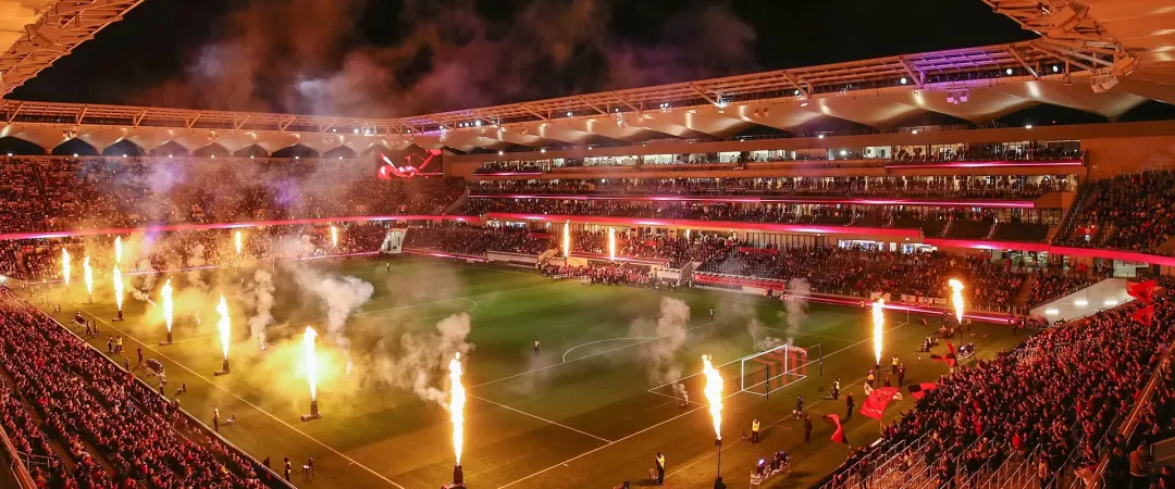 A packed soccer stadium glows under orange lights as fireworks and smoke rise around the pitch, creating a dramatic, celebratory match-night atmosphere.