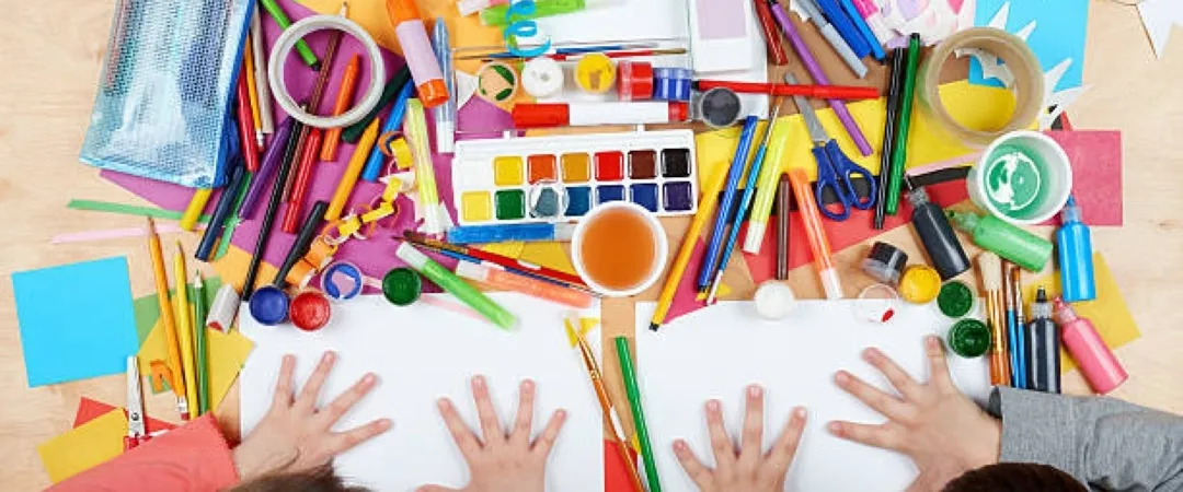 Overhead view of children reaching across blank paper amid colourful art supplies, paint, pencils, and scissors, creating a playful scene.