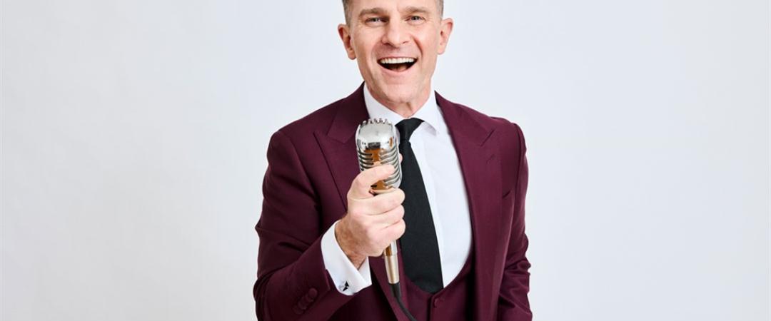 A man in a burgundy suit and black tie holds a microphone while smiling and singing against a plain white background, conveying energy and joy.