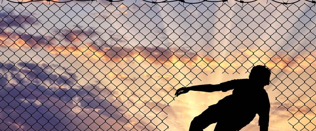 Silhouette of a person crouching next to a chain-link fence topped with barbed wire, set against a dramatic sunset sky, evoking themes of confinement.