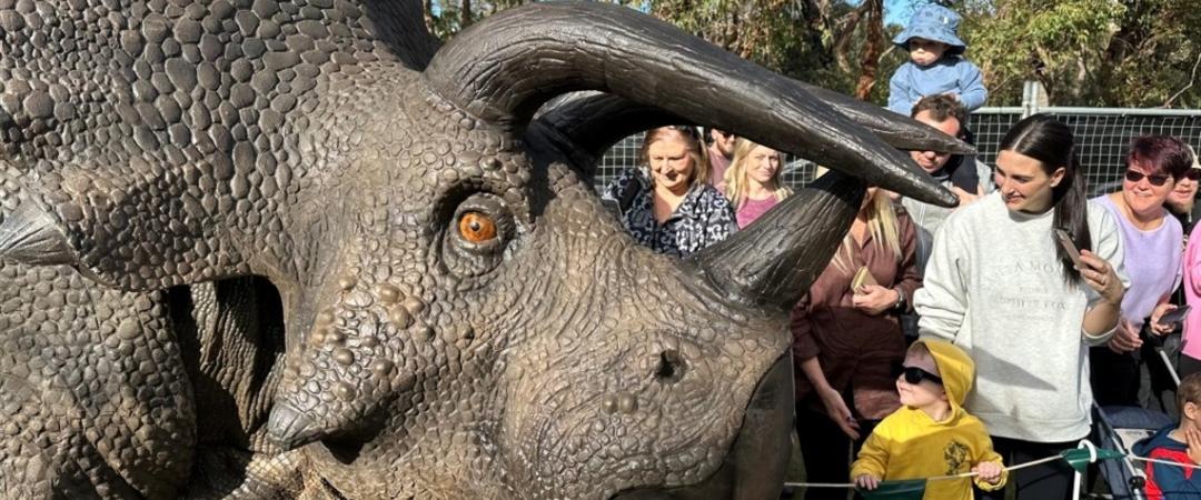 Lifelike triceratops model in a crowded park setting, surrounded by curious children and adults. The scene is lively and filled with excitement.