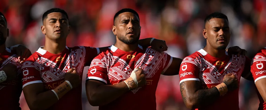 A group of rugby players in red and white jerseys stand in a row with hands on their chests, displaying focus and unity against a blurred crowd backdrop.