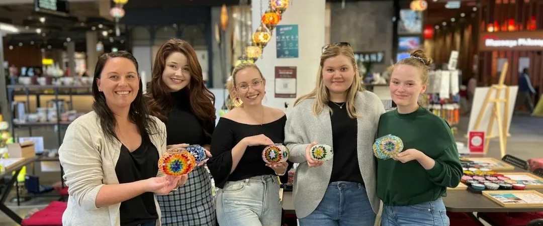 Five women smile while holding colorful mosaic coasters in a shop with decorative lighting. Their expressions convey joy and creativity.