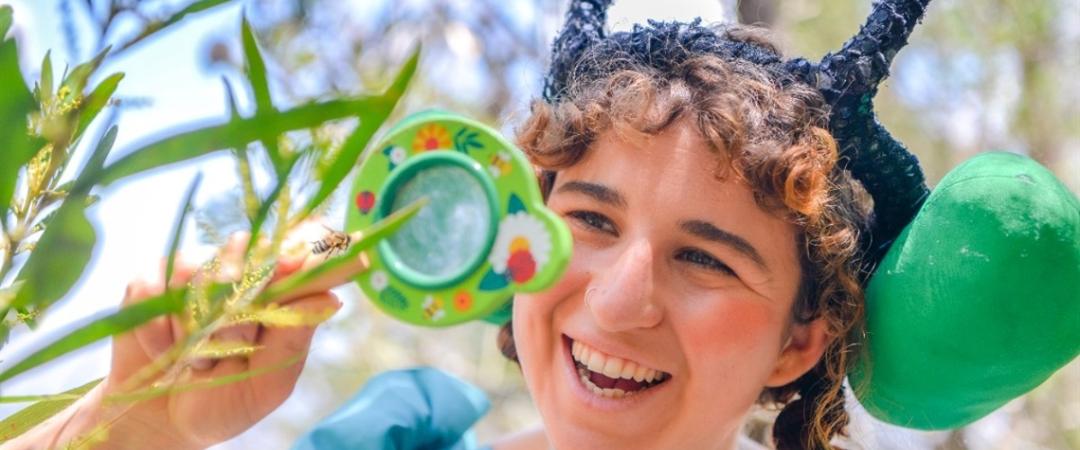 A smiling person wearing a costume with antennae examines plants through a magnifying glass in a sunny setting, conveying a sense of curiosity and playfulness.