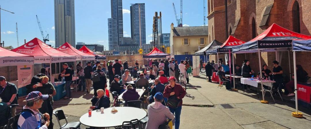 Outdoor community event with people gathered around tables. Red tents provide shade, and tall city buildings rise in the background under a clear blue sky.