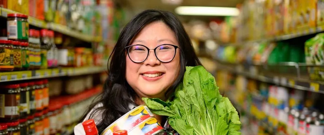 A smiling woman stands in a grocery aisle, holding canned goods, bottles of sauce, and fresh greens. Shelves filled with colorful products surround her.