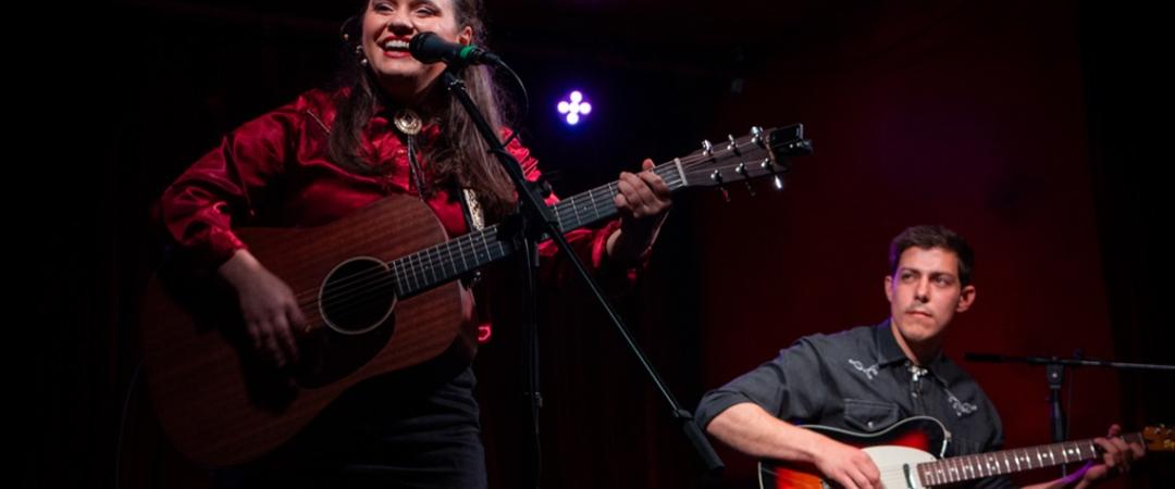 A woman in a red blouse joyfully plays an acoustic guitar and sings into a microphone on stage. Beside her, a man plays an electric guitar, adding depth to the live performance. The dimly lit setting with purple lighting creates an intimate, lively atmosphere.