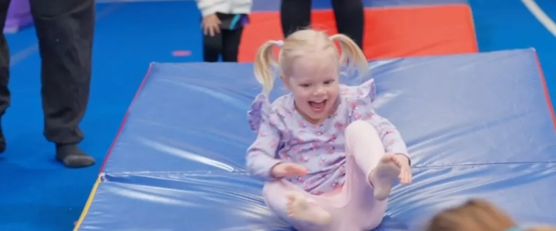 A young girl with blonde pigtails joyfully slides on a blue mat in a colorful play area, smiling widely. Other children and adults are seen in the background.