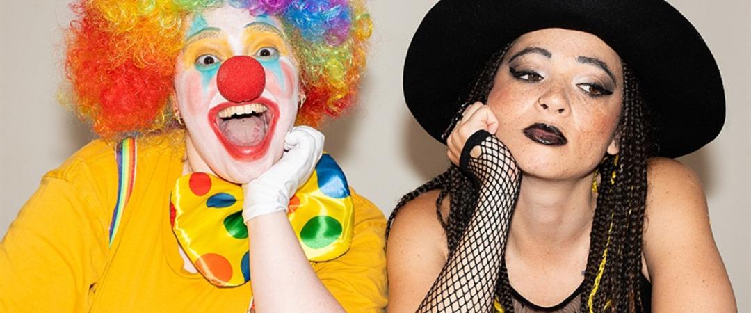 A vibrant clown with a rainbow wig and large polka-dot bow grins widely beside a contemplative woman in a black hat and fishnet sleeves, contrasting expressions.