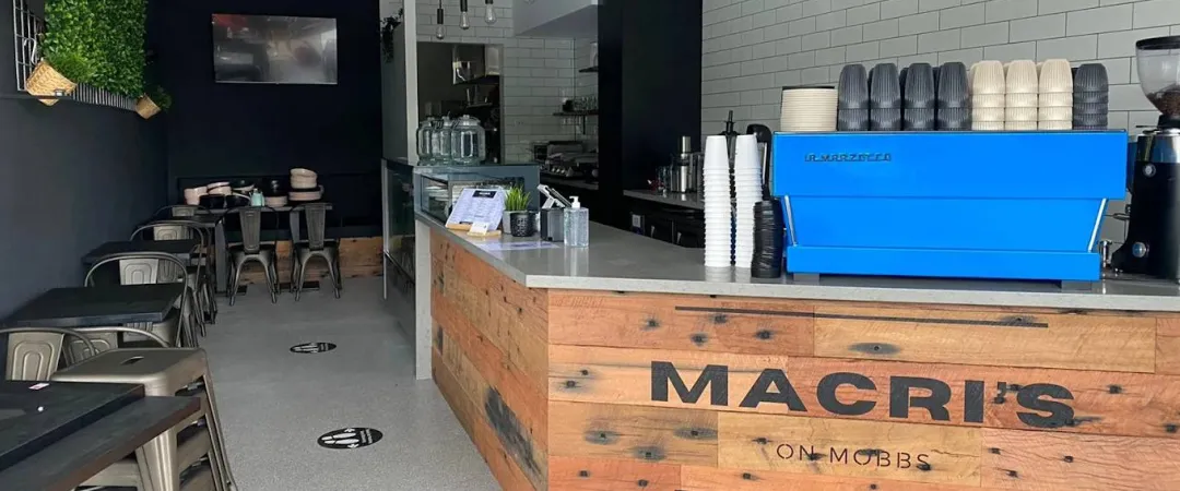 Modern café interior with white tile walls, wood counter labeled "MACRI'S," and vibrant blue espresso machine. Dark chairs line tables; pendant lights hang.