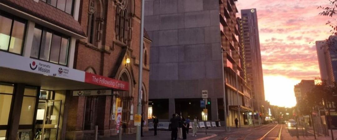 Street view at dusk with a mix of old and modern buildings under a vibrant sunset. A historic church, tram tracks, and few pedestrians are visible.