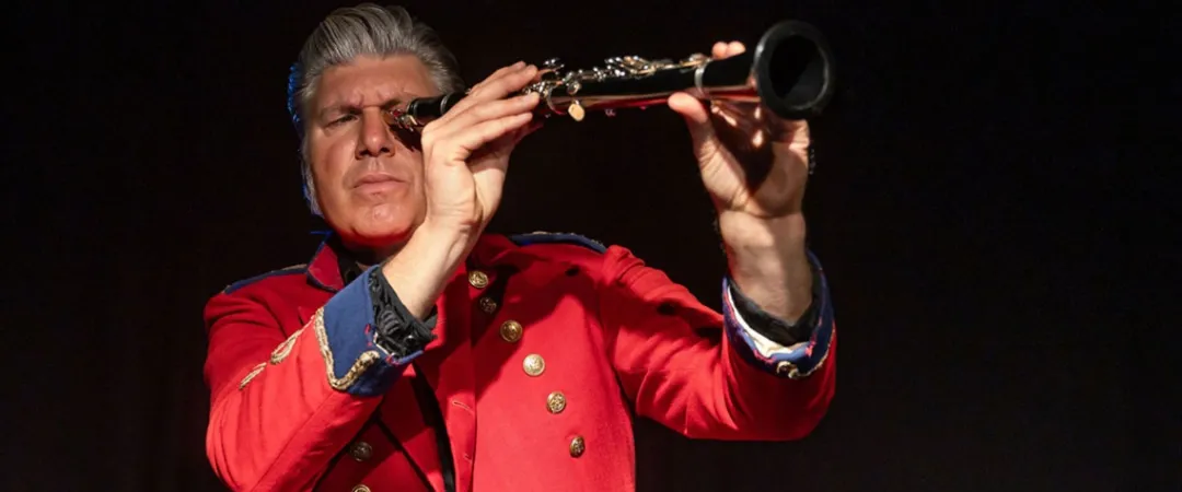 A man in a vibrant red blazer passionately plays a clarinet against a dark background, conveying dedication and concentration in his performance.