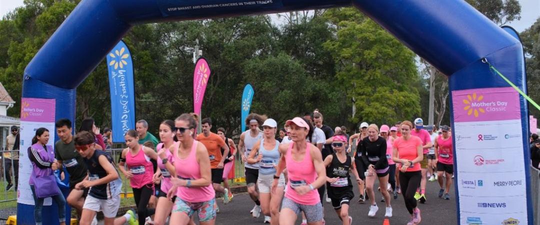 Participants in pink attire cross the starting line of a race under a blue inflatable arch. The scene is lively, with trees and banners in the background.
