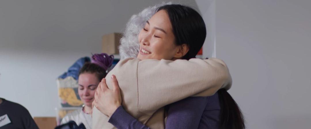 A young woman warmly hugs an older person, smiling gently, surrounded by people sorting clothes in a bright room. The mood is caring and supportive.