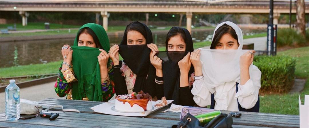 Four people sitting at an outdoor table, laughing and covering their faces with colourful scarves. A cake and a camera are on the table, conveying a joyful and playful mood.