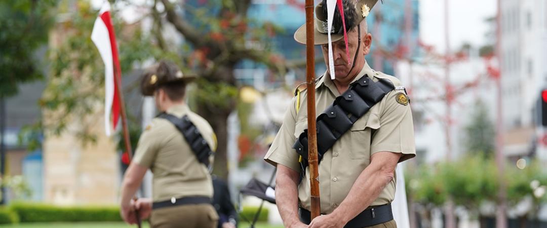 Two uniformed individuals in ceremonial attire stand at attention near a flagpole in a green, urban park, conveying a solemn and respectful atmosphere.