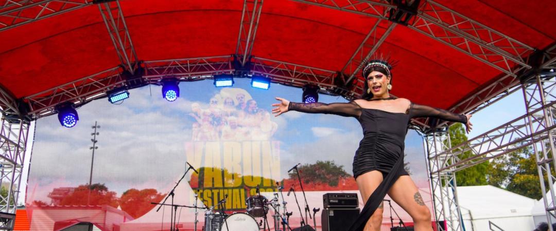 Drag performer in a black dress and headpiece on a vibrant outdoor stage with a red canopy. Background shows musical instruments and festival banner. Energetic atmosphere.