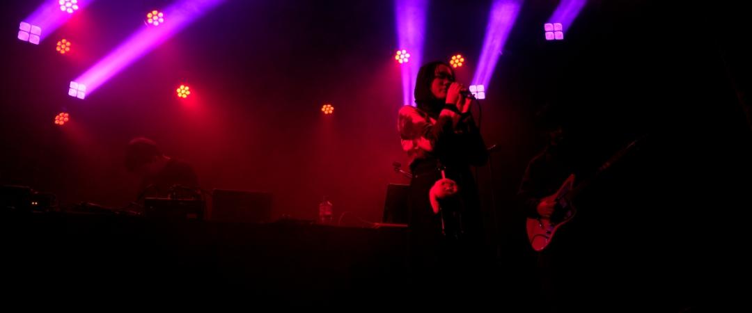 A singer performing on stage with vibrant purple and pink lights illuminating the background. A guitarist plays beside them, creating an energetic atmosphere.