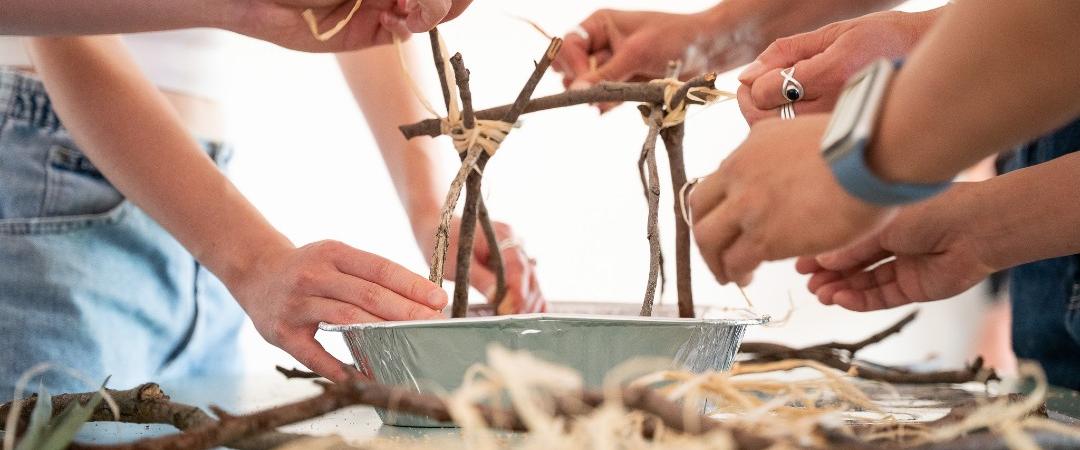 Several people work together to build a small structure from sticks and twine over a metal tray, with more sticks and materials scattered on the table.