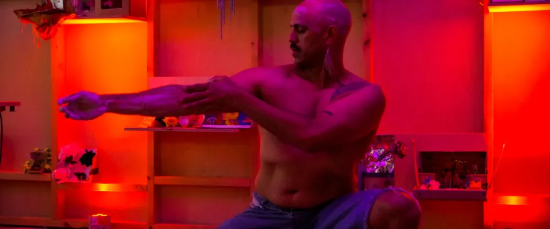 A shirtless man with a shaved head and mustache kneels indoors, pulling on his left arm, surrounded by shelves lit with red and purple lighting.