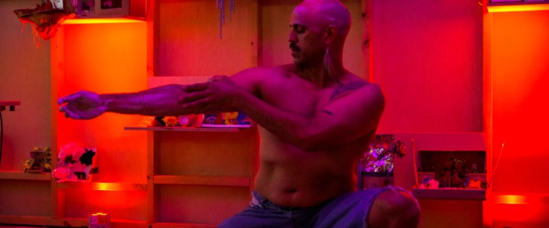 A shirtless man with a shaved head and mustache kneels indoors, pulling on his left arm, surrounded by shelves lit with red and purple lighting.