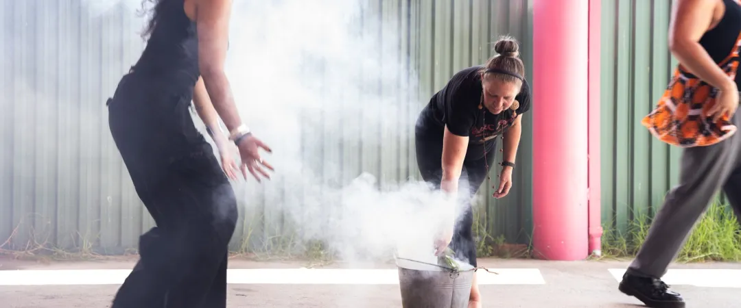 Two people stand near a metal bucket producing smoke, with one person bending over the bucket and another standing beside, outdoors under a covered area.