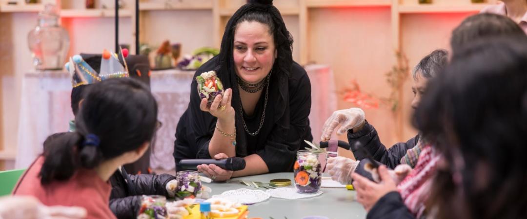 A woman smiles while holding a jar with colorful contents, engaged with a group crafting at a table. The setting is warm and creative, decorated with plants.