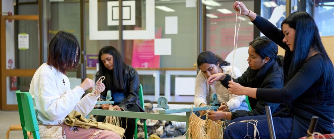 Five people sit in a circle weaving with long strands of natural fiber in an indoor setting, surrounded by weaving materials on the floor.