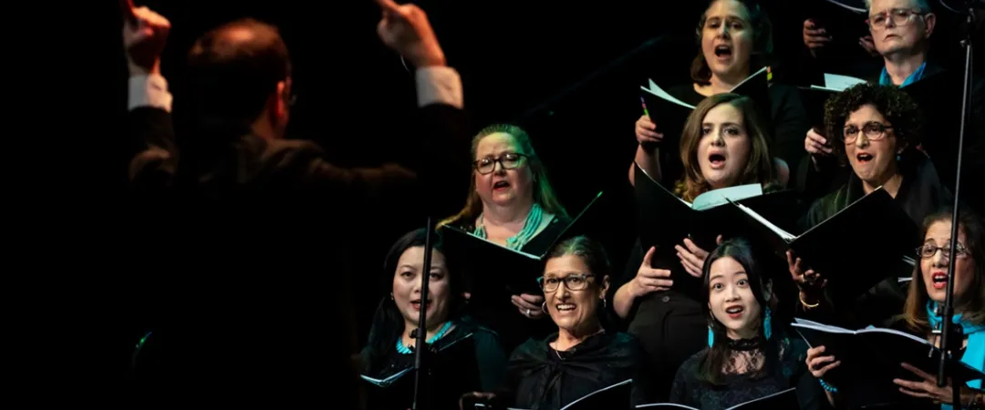 A choir of women dressed in black sings passionately, expressions full of emotion, against a dark background. Their hands are raised, conveying depth and energy.