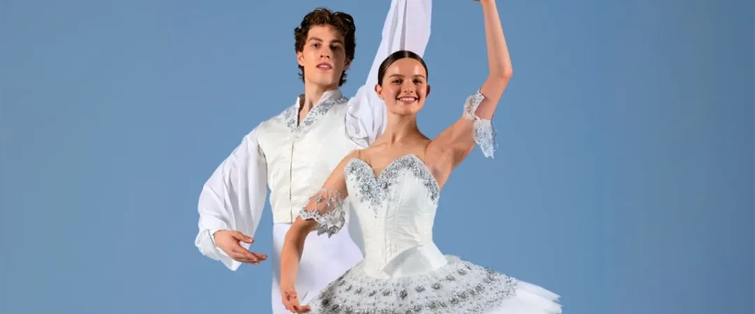 A male and female ballet dancer pose gracefully against a blue background. The woman, in a white tutu, raises an arm; the man stands elegantly beside her in matching attire.