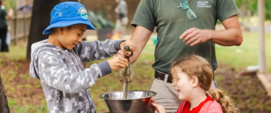 A man in a hat helps two smiling children operate an old-fashioned hand crank over a bowl outdoors. The scene is cheerful, set in a park with trees.
