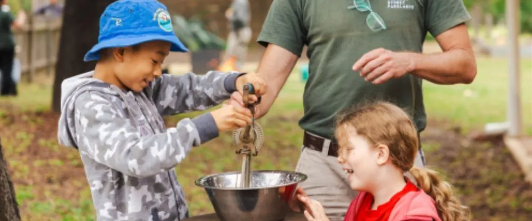 A man in a hat helps two smiling children operate an old-fashioned hand crank over a bowl outdoors. The scene is cheerful, set in a park with trees.