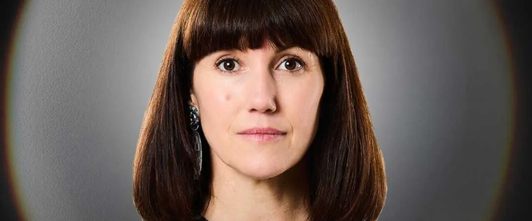 A woman with straight, dark brown hair and bangs, wearing a black top, stares seriously at the camera. The background is a blurred gray gradient.