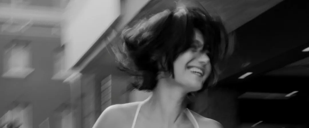 Black-and-white, motion-blurred portrait of a woman in a sleeveless top turning with a joyful smile, her hair in motion against an urban backdrop.