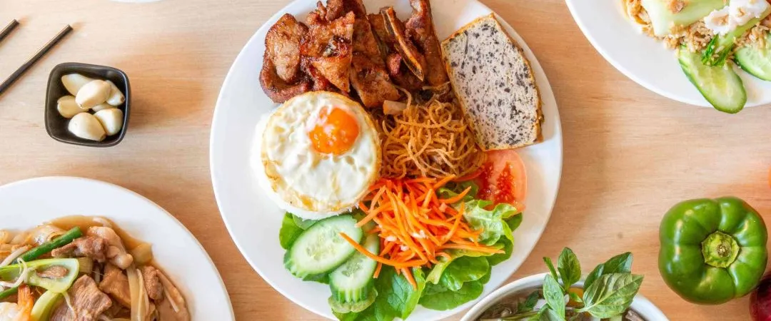 A top-down view of a wooden table featuring a central plate of pork, egg, and rice surrounded by bowls of noodle soup, fried rice, and fresh produce.