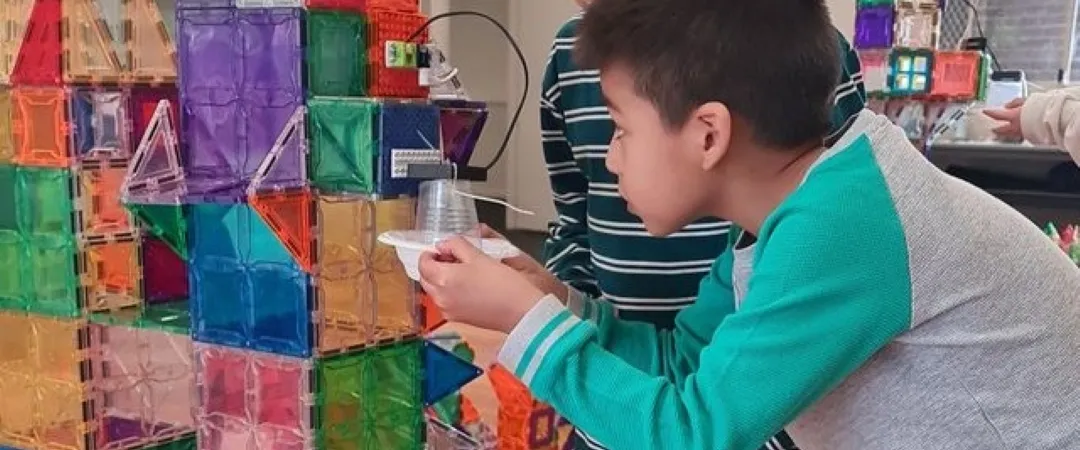 Two boys collaborate on a colorful magnetic-tile structure, examining a small piece indoors; the scene conveys curiosity, teamwork, and learning.