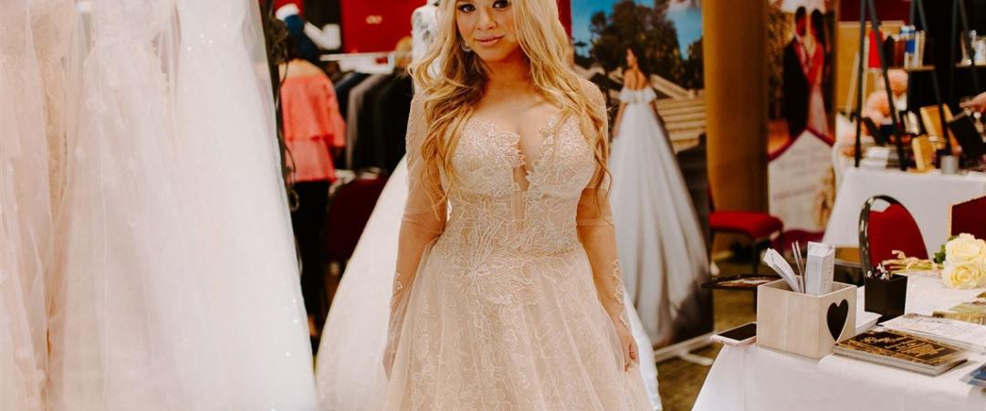 A smiling woman in a bridal shop wears an elegant, beaded wedding gown with a flowing skirt. Surrounding are racks of white dresses and decor.