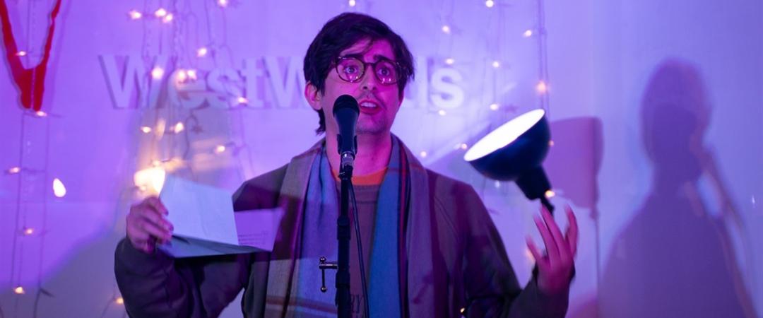 A person stands by a microphone, holding a paper and a lamp, surrounded by twinkling fairy lights. The scene is vibrant with creative energy.