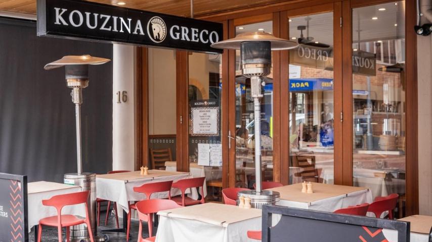Tables with red checker table clothes sitting in front of restaurant with name overhead 'Kouzina Greco'