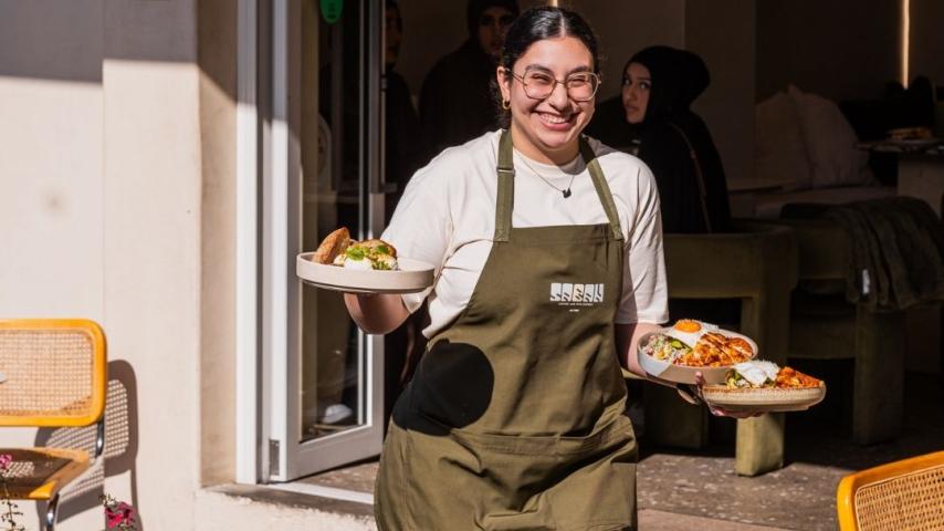 Women with big smile, wearing white shirt and green apron holding plates of food 
