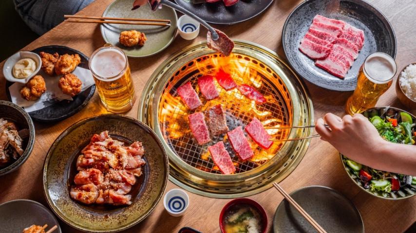 Table with grill in the middle and hands putting raw meat to cook. 