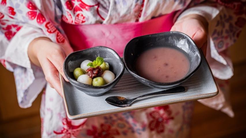 Person in pink kimono holding platter with bowl of purple liquid and small green mochi balls 
