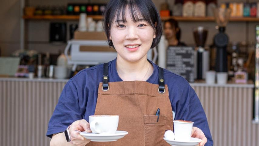 Asian looking woman wearing brown apron holding 2 cups of coffees in white mugs