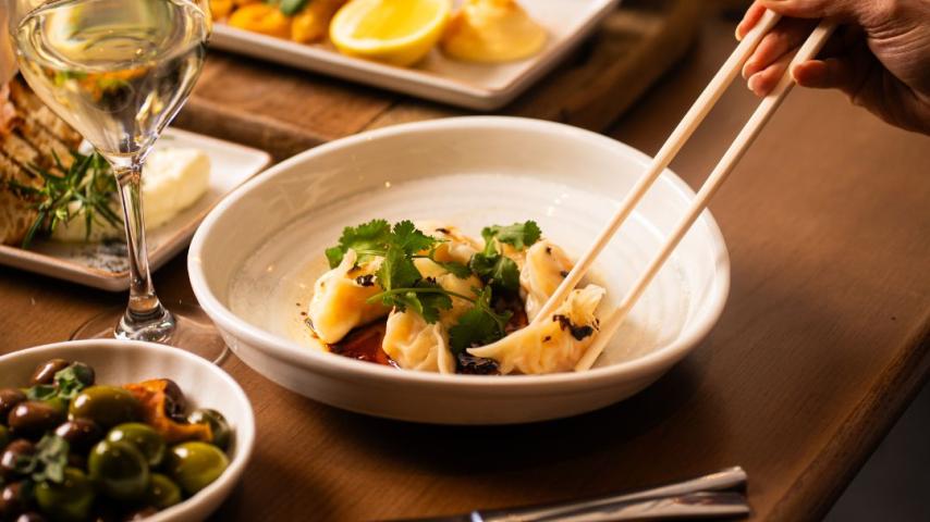 A hand uses chopsticks to pick up dumplings garnished with cilantro in a white bowl. Surrounding dishes include olives, wine, and lemon slices, creating a cozy dining scene.