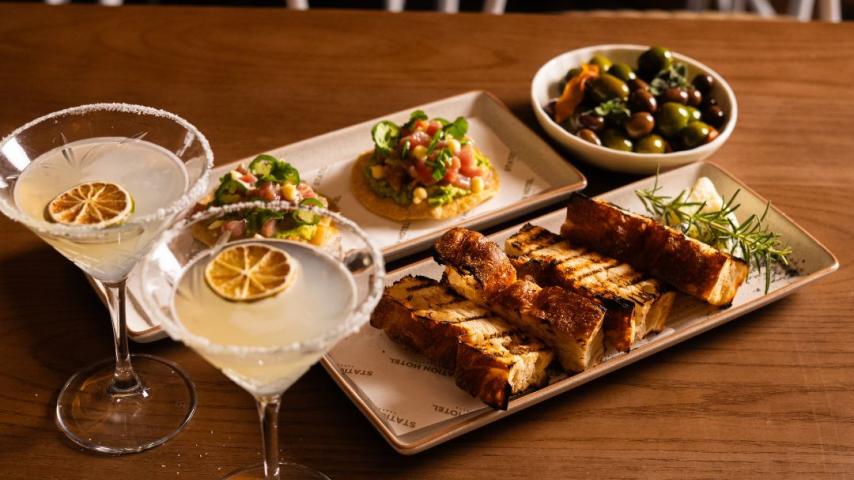 Two martinis with dried lemon slices, grilled bread, a taco with greens, and a bowl of mixed olives on a wooden table, creating a cozy and elegant dining atmosphere.